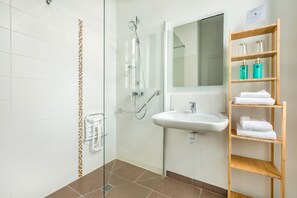 Un baño con ducha envuelta en vidrio, un lavamanos blanco, un espejo y un estante de madera con toallas.