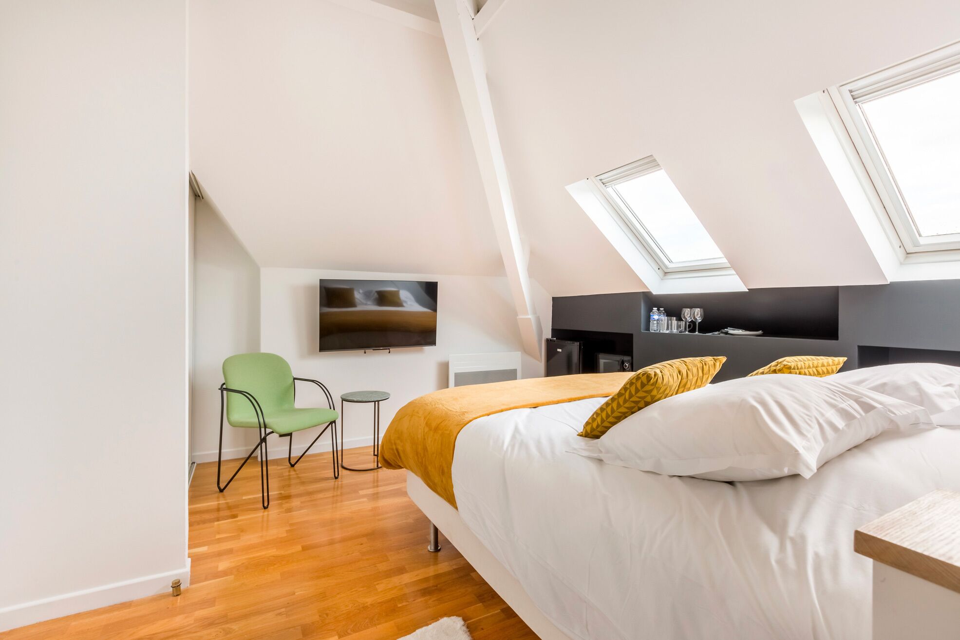 Un dormitorio moderno con una cama grande, una silla verde, una mesita pequeña, un televisor y un minibar.