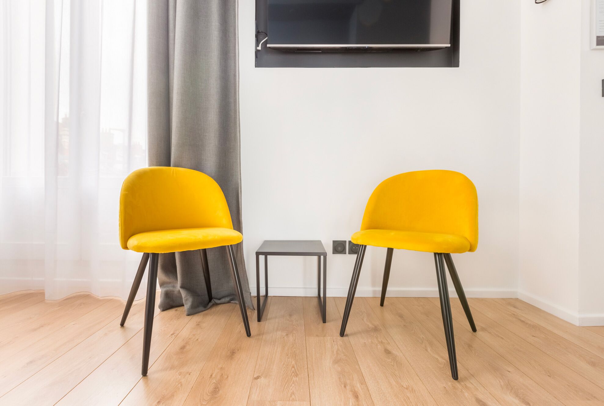 Two yellow chairs, a small table, and a TV mounted on the wall in a room with wooden flooring and a grey curtain.