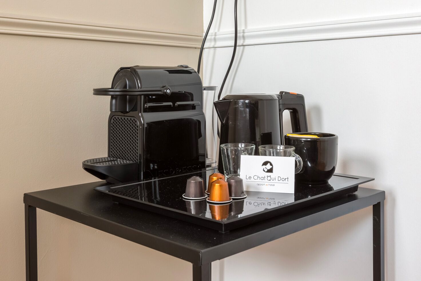A coffee station with a coffee maker, a kettle, a mug, and a sign for "Le Chat Qui Dort" hotel.