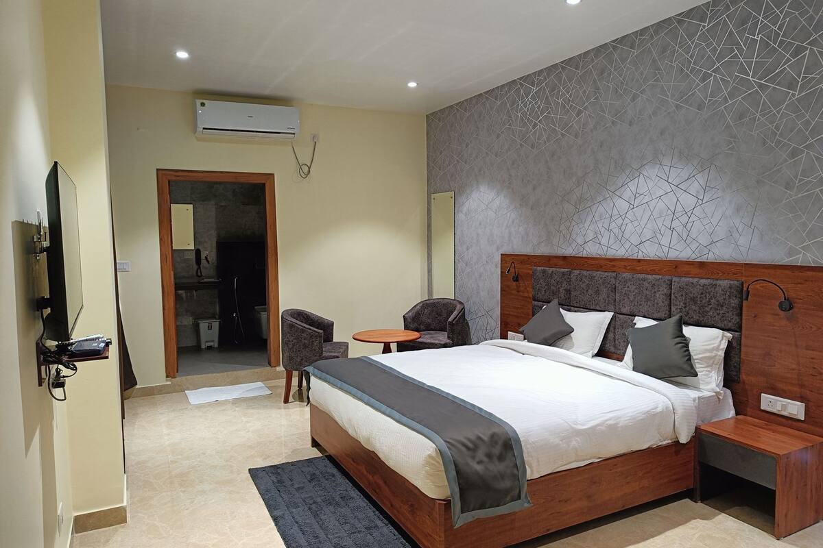 A modern hotel room with a wooden bed, two armchairs, a small round table, and a built-in air conditioning unit.