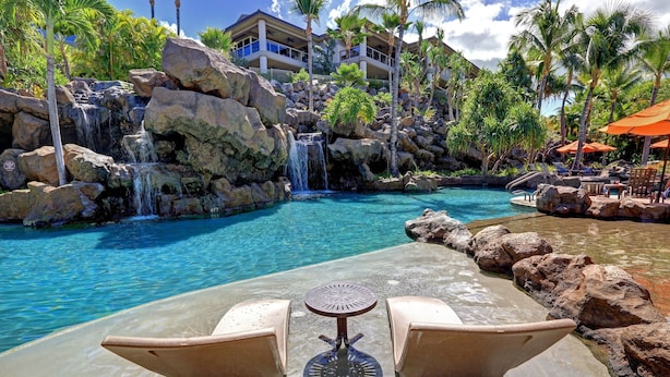A poolside area with a waterfall, lounge chairs, and a view of a resort building. - Ho'olei Garden View by Coldwell Banker Island Vacation (Kihei)