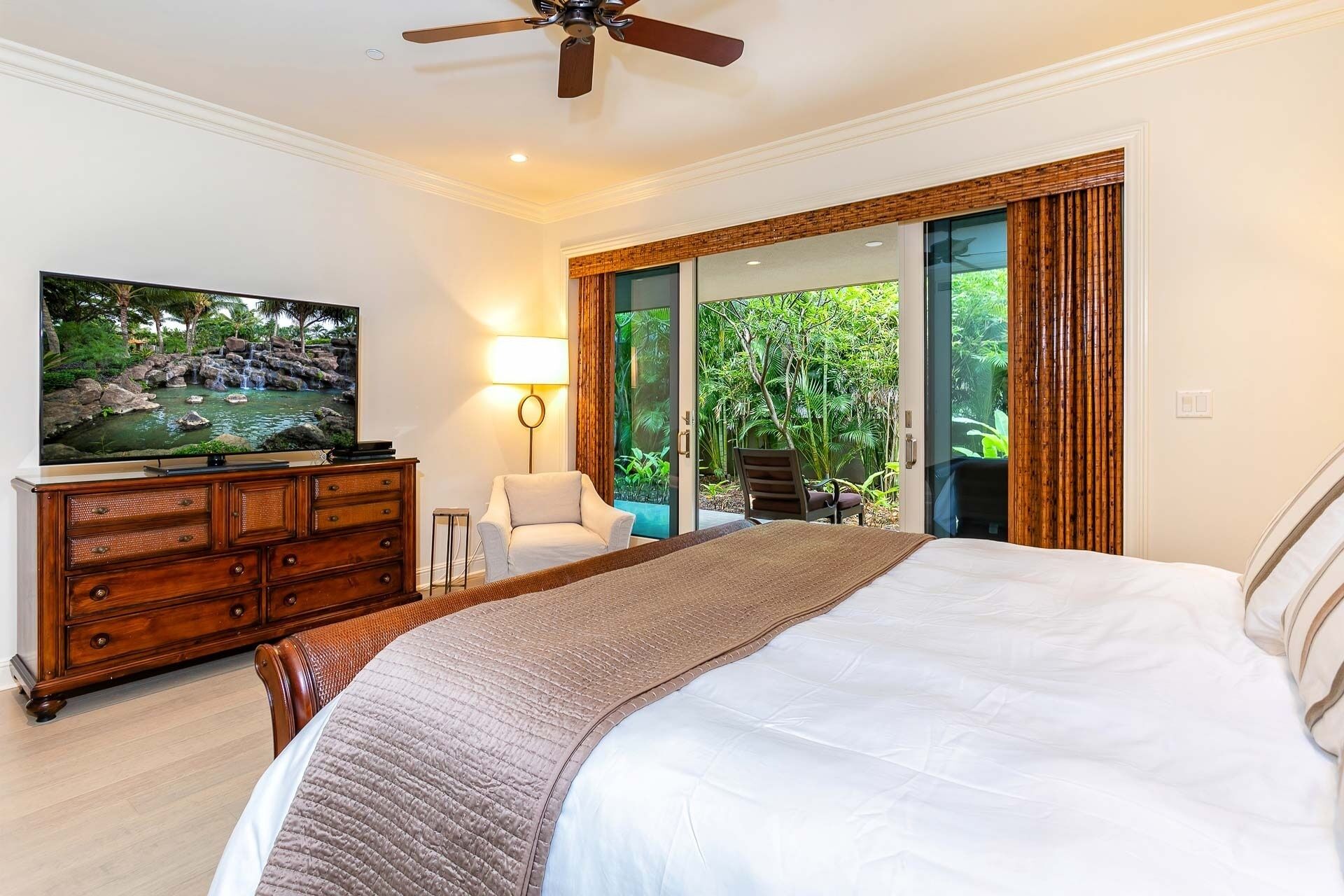 A bedroom with a bed, a TV on a wooden dresser, a chair, and a view of greenery through a glass door.