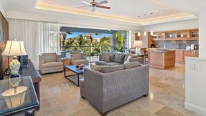 A spacious living area with a sofa set, a coffee table, and a glass-top table. There is a kitchen area with wooden cabinets and a large island. The room has a ceiling fan and large windows with a view of palm trees. - Ho'olei Garden View by Coldwell Banker Island Vacation (Kihei)