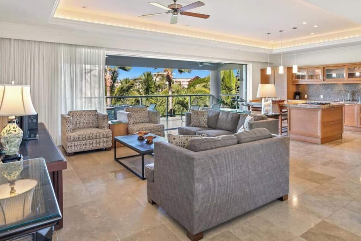Amplia sala de estar con un juego de sofá, una mesa de centro y una mesa con superficie de vidrio. Cuenta con una zona de cocina con armarios de madera y una isla grande. La habitación tiene un ventilador de techo y ventanas amplias con vistas a palmeras.