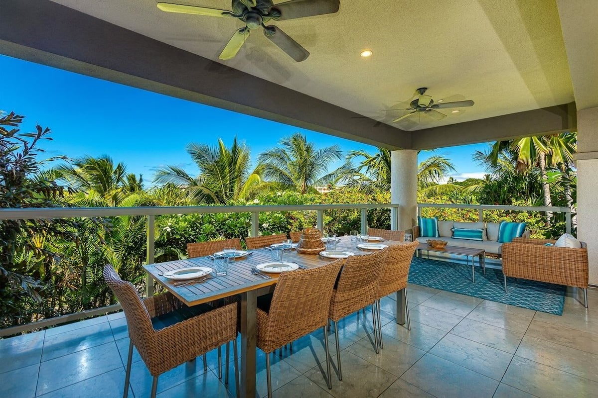 Un área de comedor al aire libre con una mesa preparada para cuatro personas, rodeada de sillas de mimbre, con vistas a un exuberante jardín tropical.