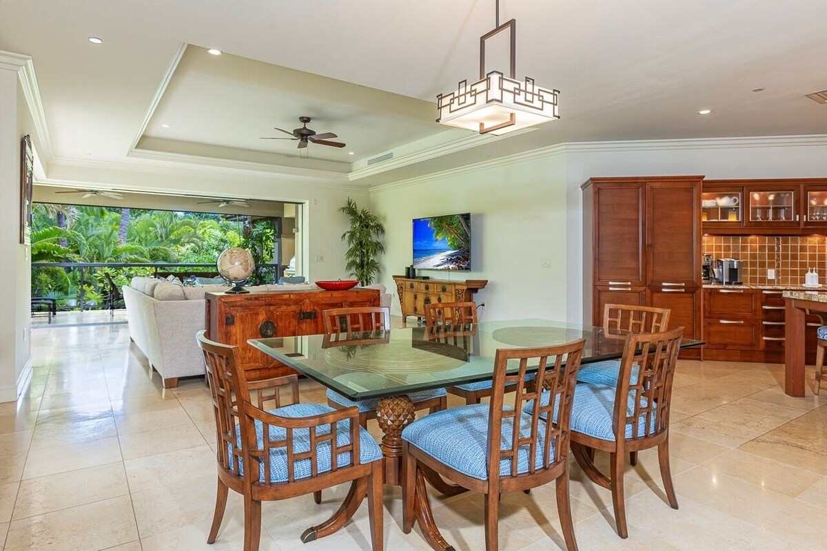 A spacious dining area with a glass-top table and wooden chairs, a ceiling fan, and a view of lush greenery outside.