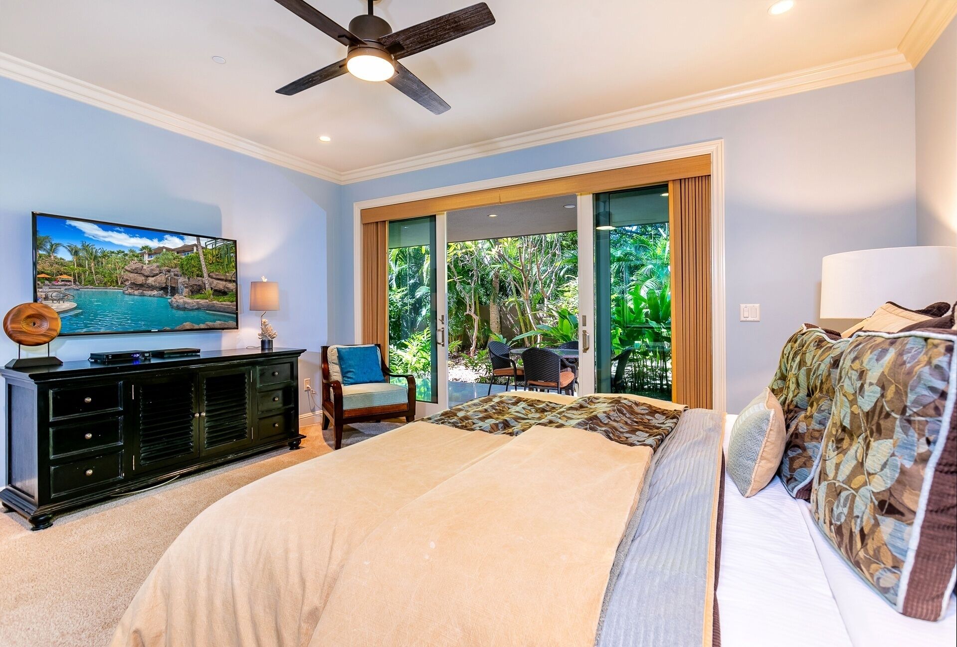 A bedroom with a bed, a TV, a ceiling fan, and a view of a tropical landscape through the sliding glass door.