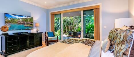 A bedroom with a bed, a TV, a ceiling fan, and a view of a tropical landscape through the sliding glass door.