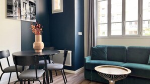 Een moderne woonkamer met een donkerblauwe accentmuur, een blauwgroene bank, een ronde salontafel en een eethoek met een kleine tafel en stoelen.