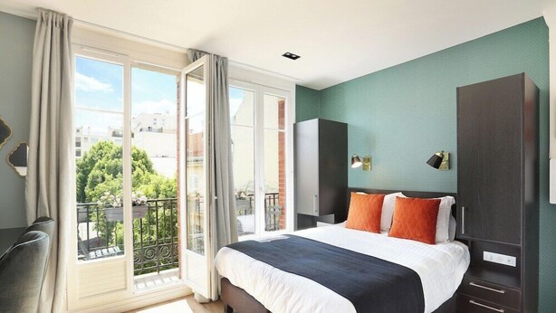 Een slaapkamer met een groot bed, een kledingkast, een bureau en een balkon met uitzicht op gebouwen en bomen.