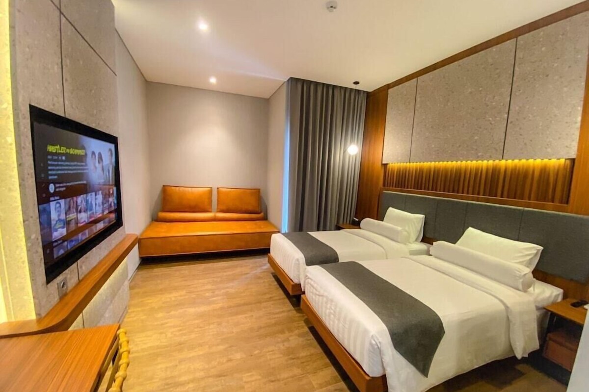 Habitación de hotel con televisión montada en la pared, un sofá, dos camas con cabeceros y suelo de madera.