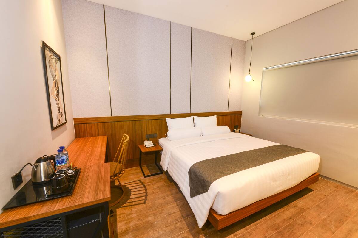 A hotel room with a bed, bedside table, and a desk with a kettle and water bottle.