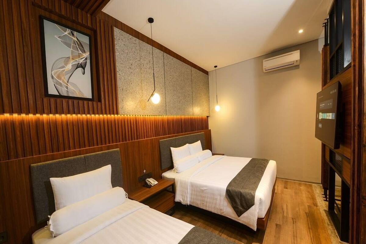 A hotel room with two beds, wooden paneling, a modern light fixture, an air conditioning unit, and a television.