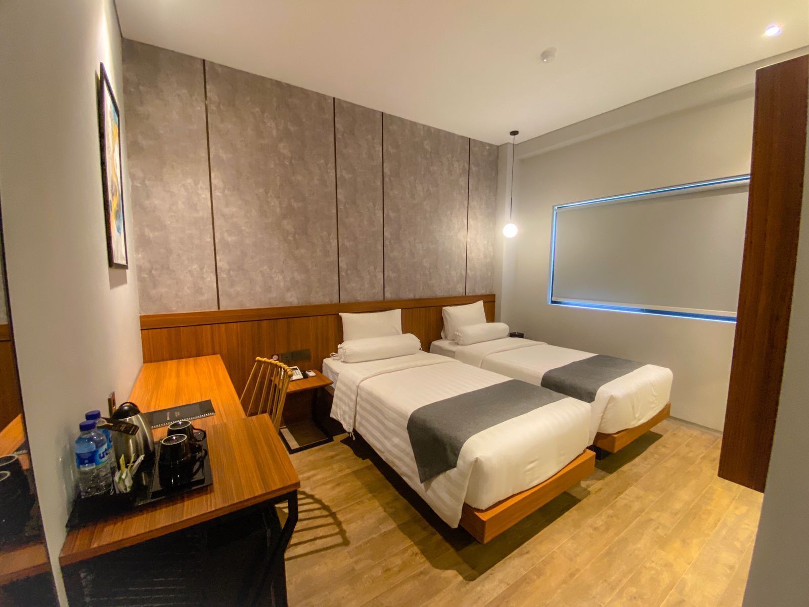 A modern hotel room with two single beds, a wooden desk with a chair, a small table with a kettle and cups, and a large window with a view.