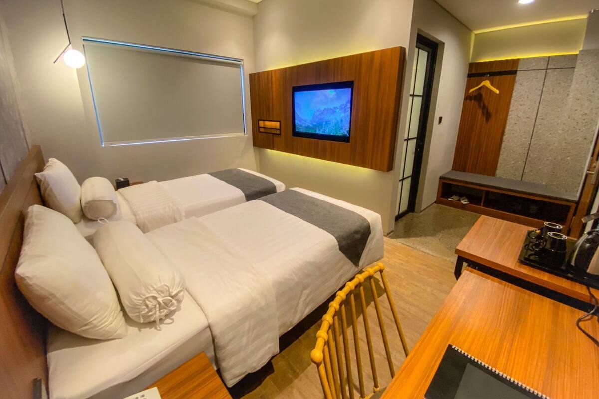A hotel room with two beds, a TV mounted on the wall, a wooden desk, and a bench.