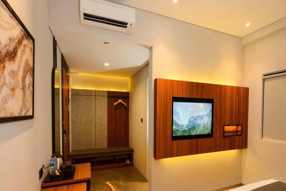 A hotel room with a bed, a TV mounted on the wall, a desk with a chair, a mirror, and an air conditioning unit.