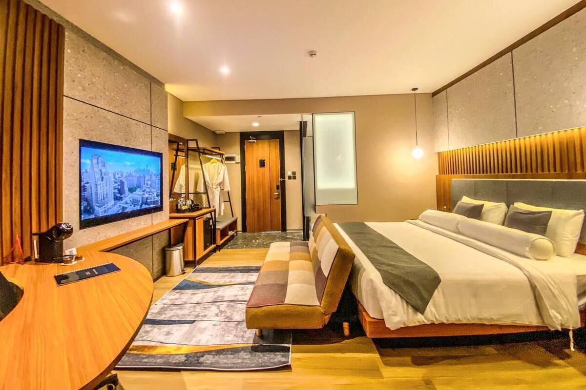 A modern hotel room with a large bed, a desk with a flat-screen TV, a seating area with a sofa, and a large window.