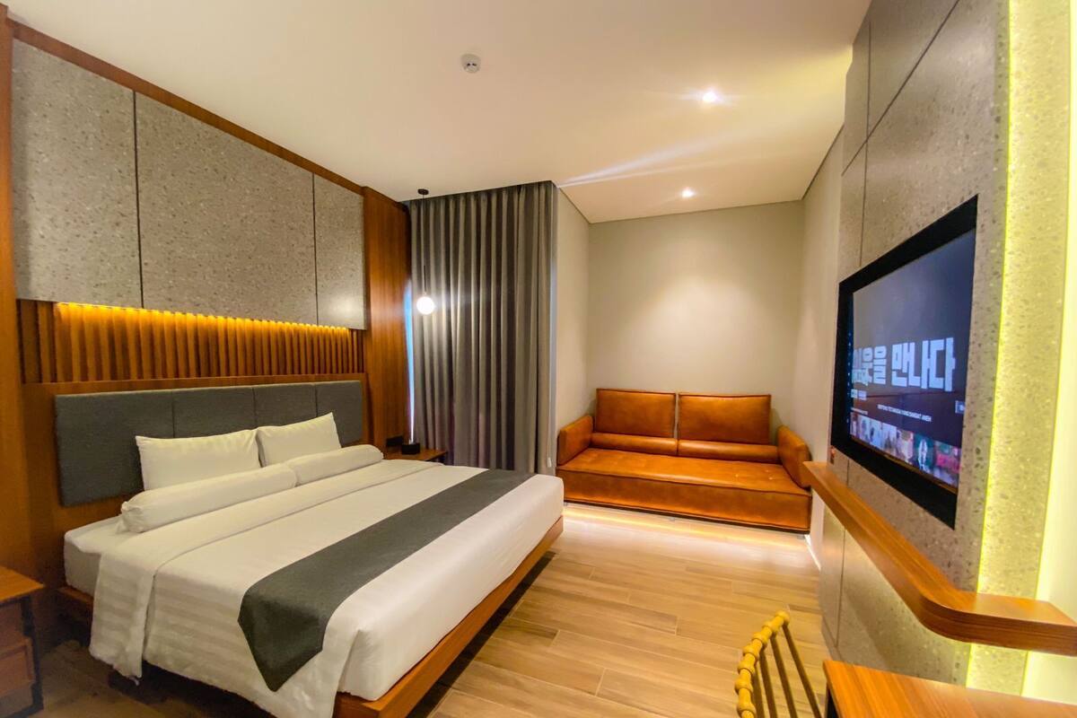 A modern hotel room with a large bed, a sofa, a TV mounted on the wall, and a wooden bench.