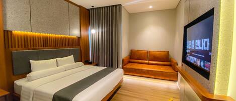A modern hotel room with a large bed, a sofa, a TV mounted on the wall, and a wooden bench.