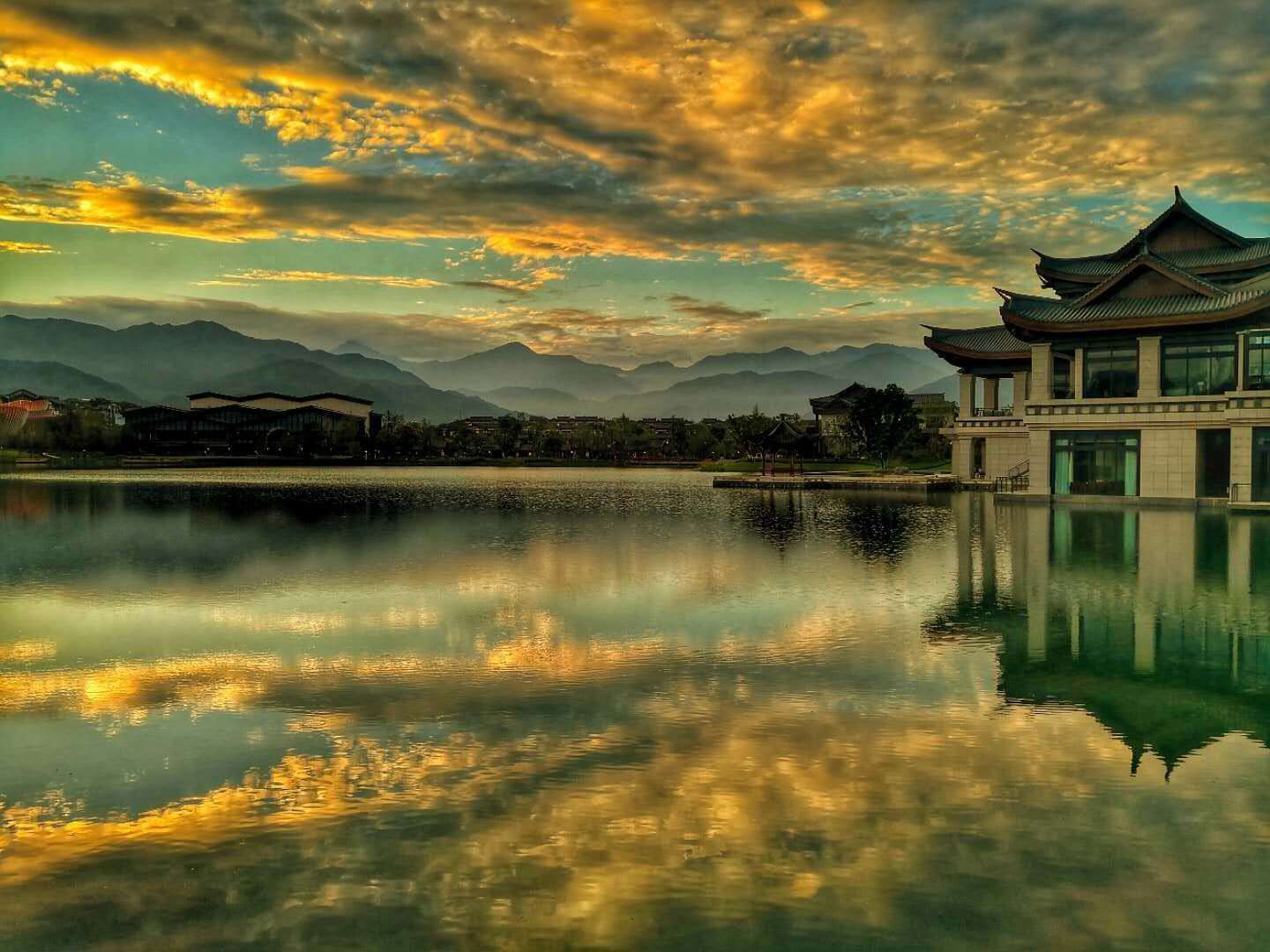 A traditional building with a reflection in a calm body of water, surrounded by mountains and a cloudy sky.