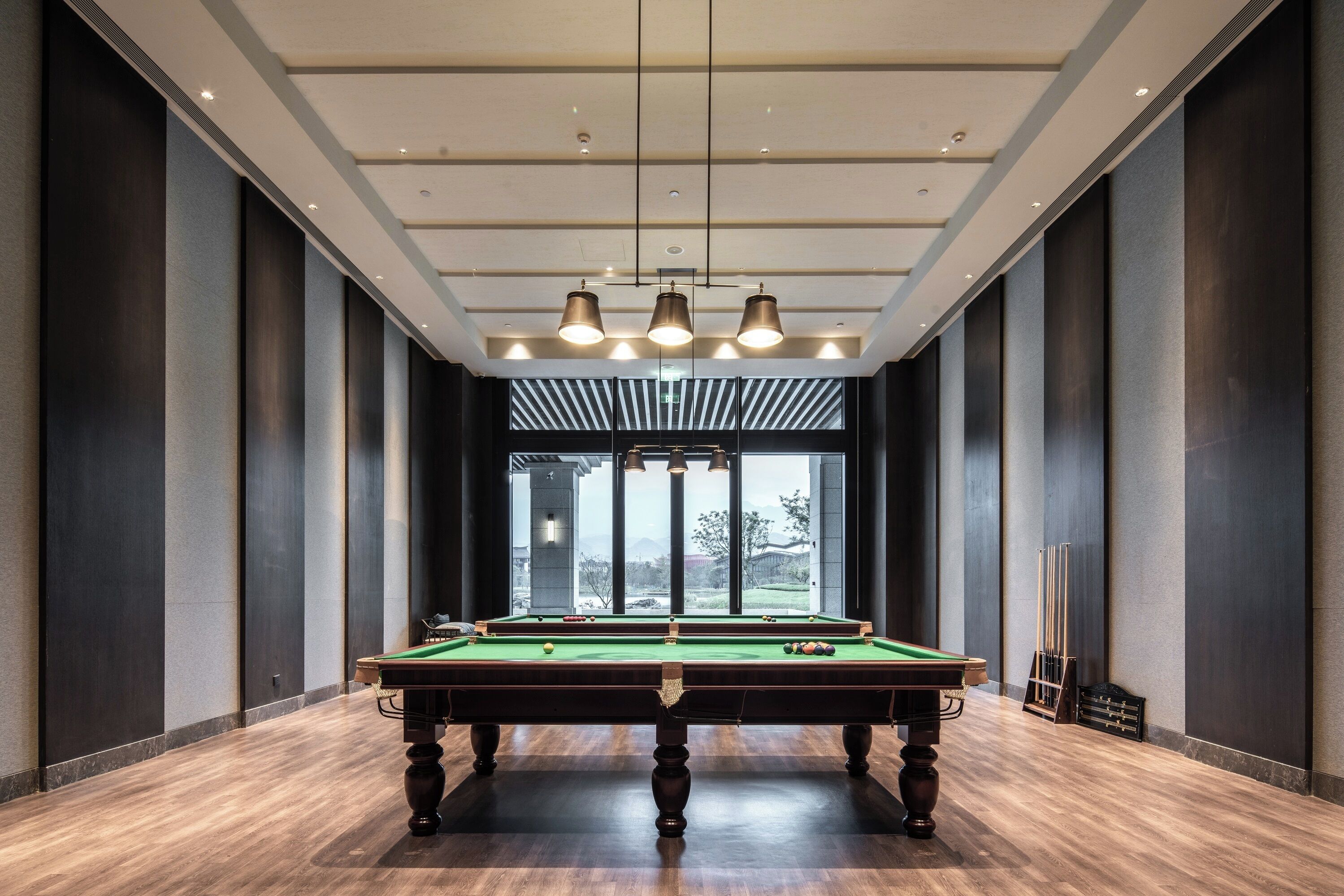 A pool table in a room with dark walls and a wooden floor, with a view of trees and buildings outside through large windows.