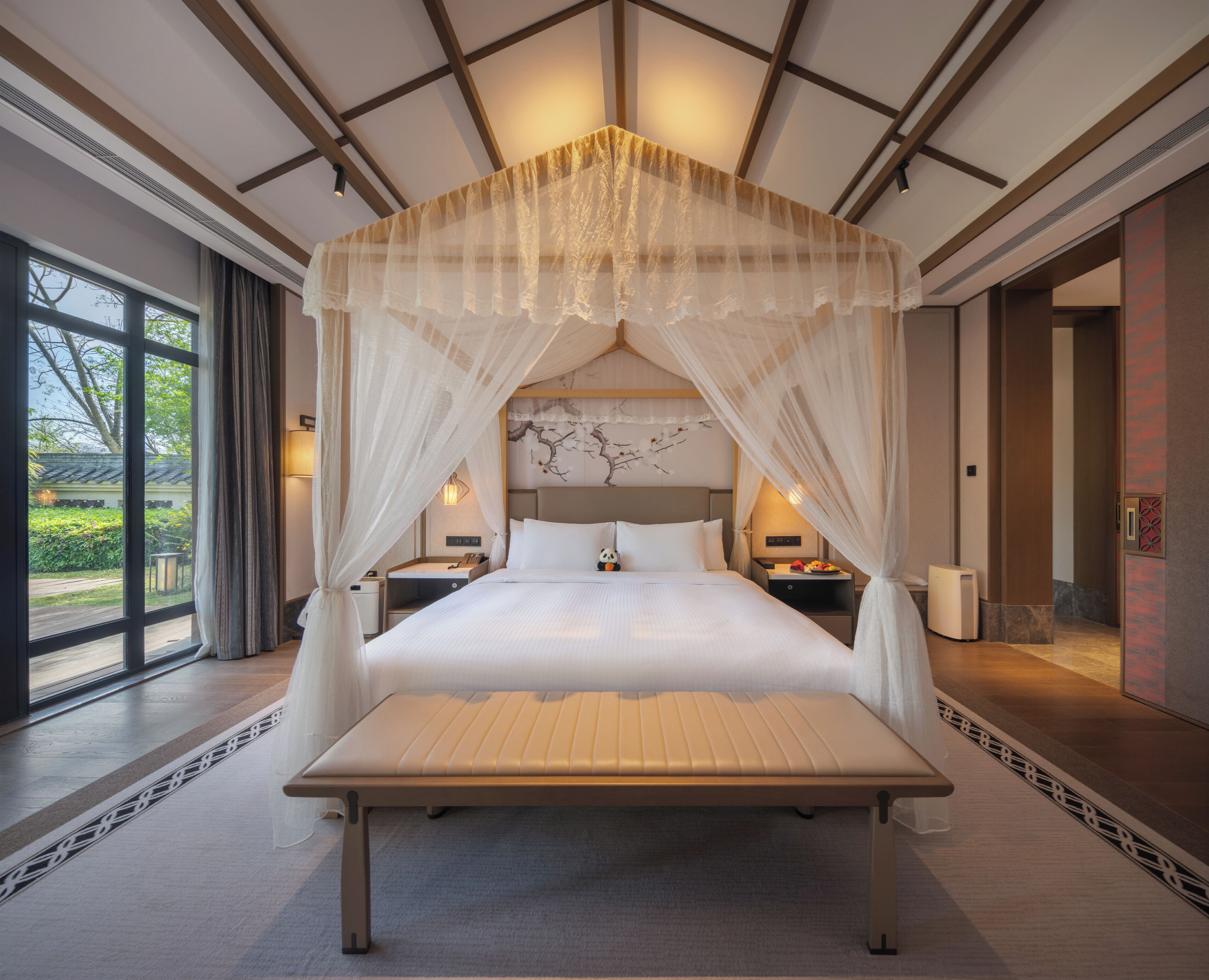 A large bed with a canopy, bedside tables, a nightstand, and a view of greenery outside.