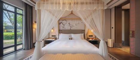 A large bed with a canopy, bedside tables, a nightstand, and a view of greenery outside.