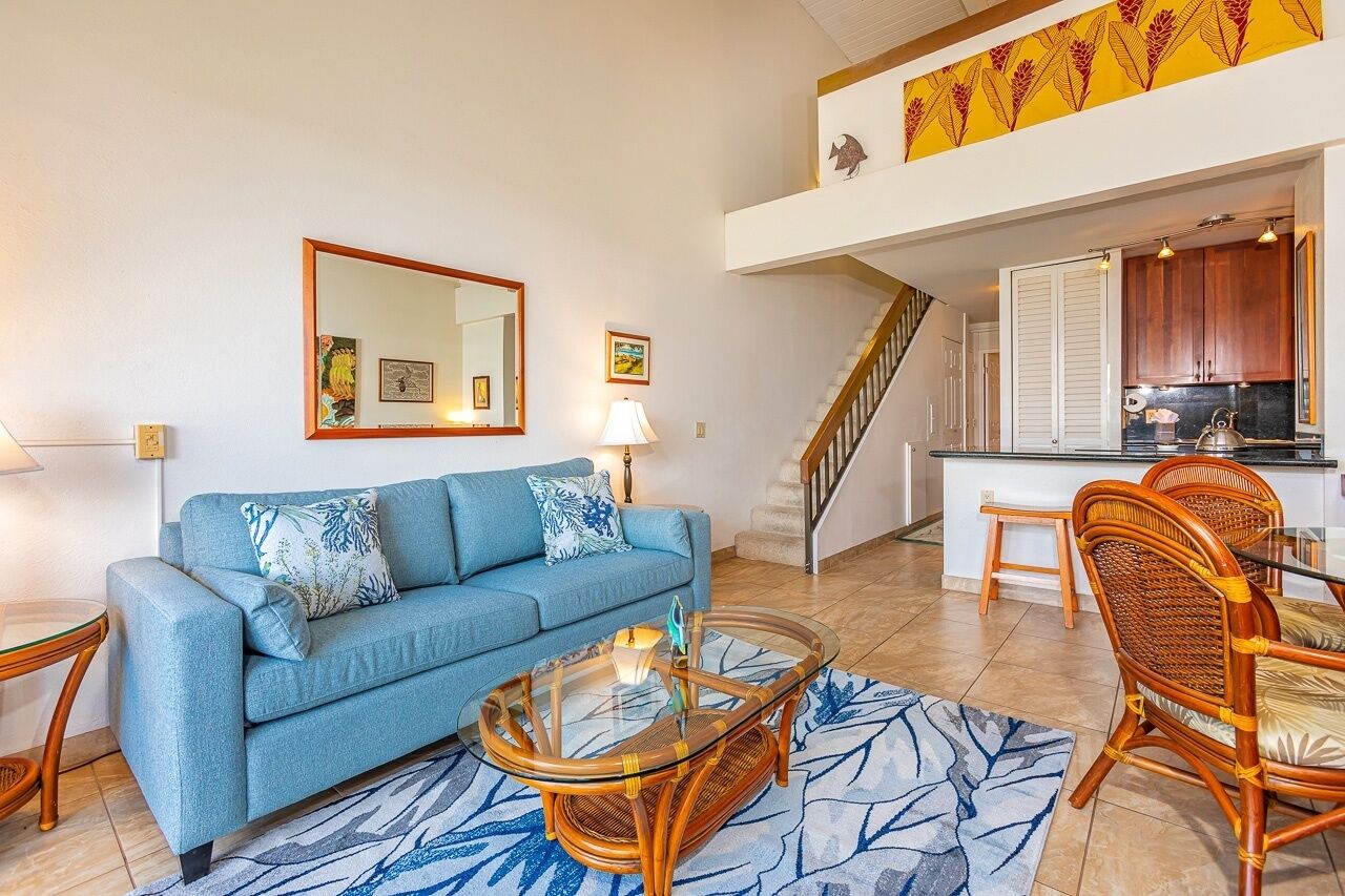 Un salon avec un canapé bleu, une table basse en verre, des fauteuils en osier, un escalier et un coin cuisine.