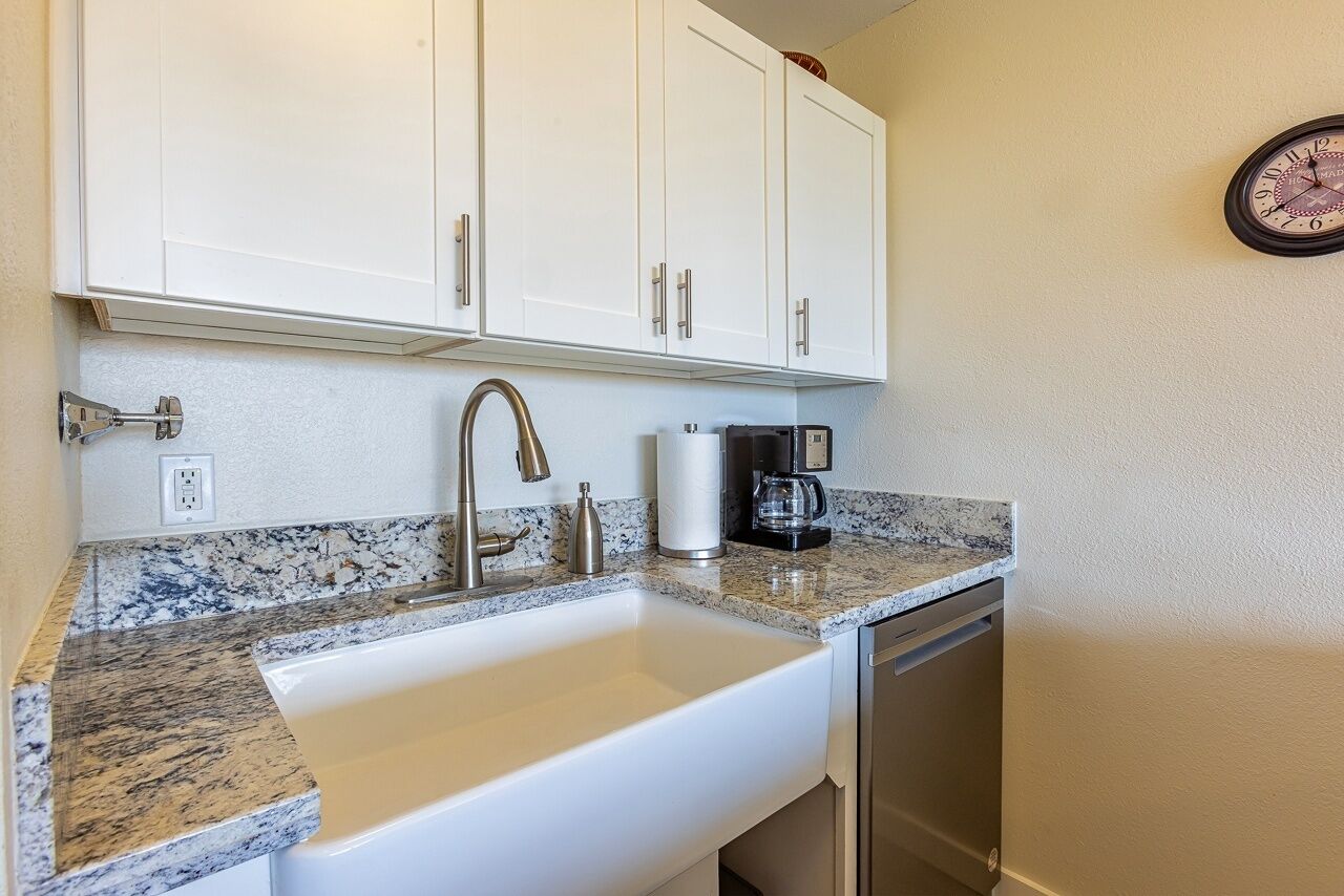 A kitchen with white cabinets, a stainless steel sink, a granite countertop, a coffee maker, and a clock on the wall.