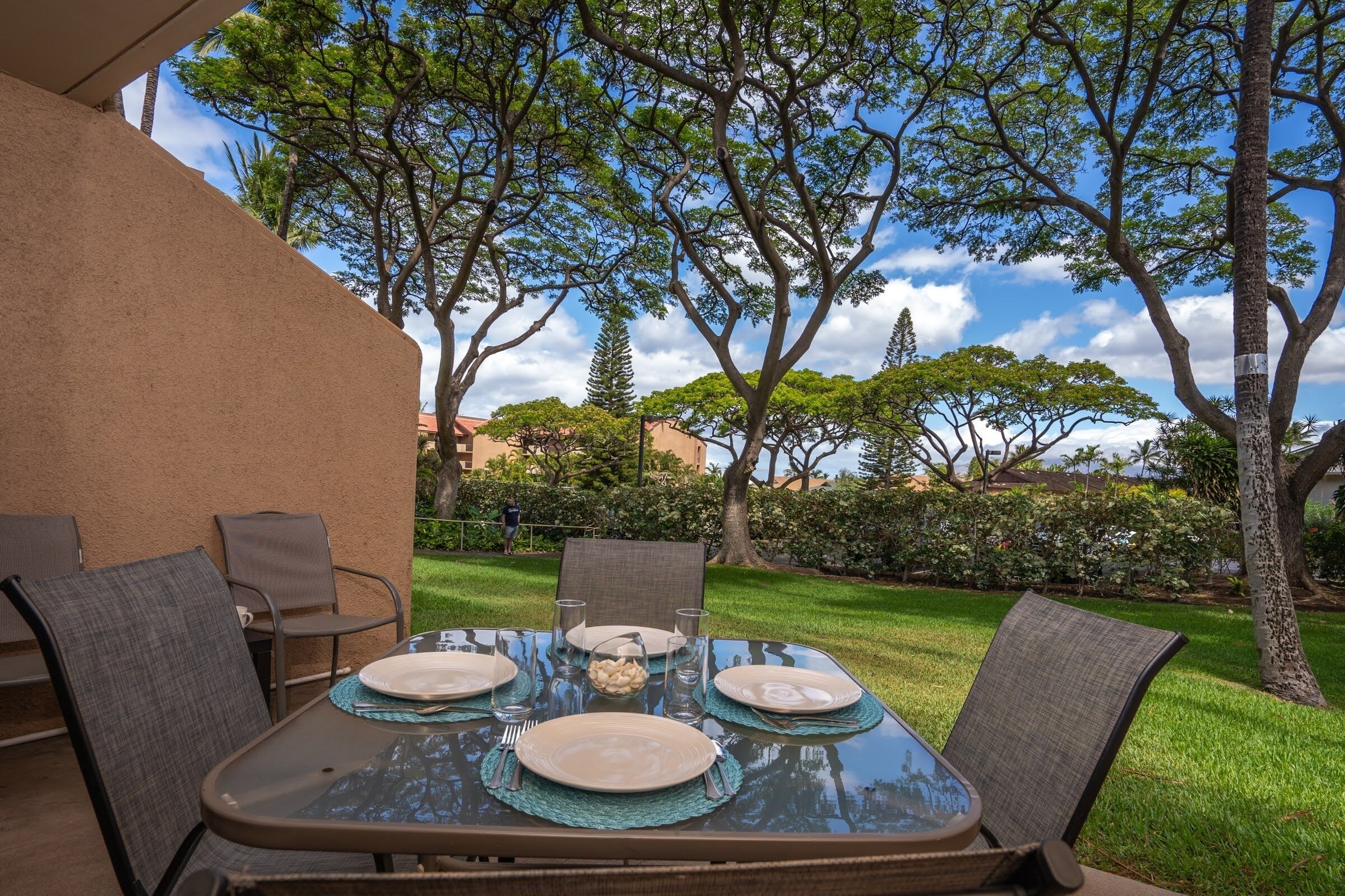 Un espace extérieur aménagé pour les repas, avec une table en verre pour quatre personnes, entourée de chaises, dans un jardin paysager agrémenté d’arbres et avec un bâtiment en arrière-plan.