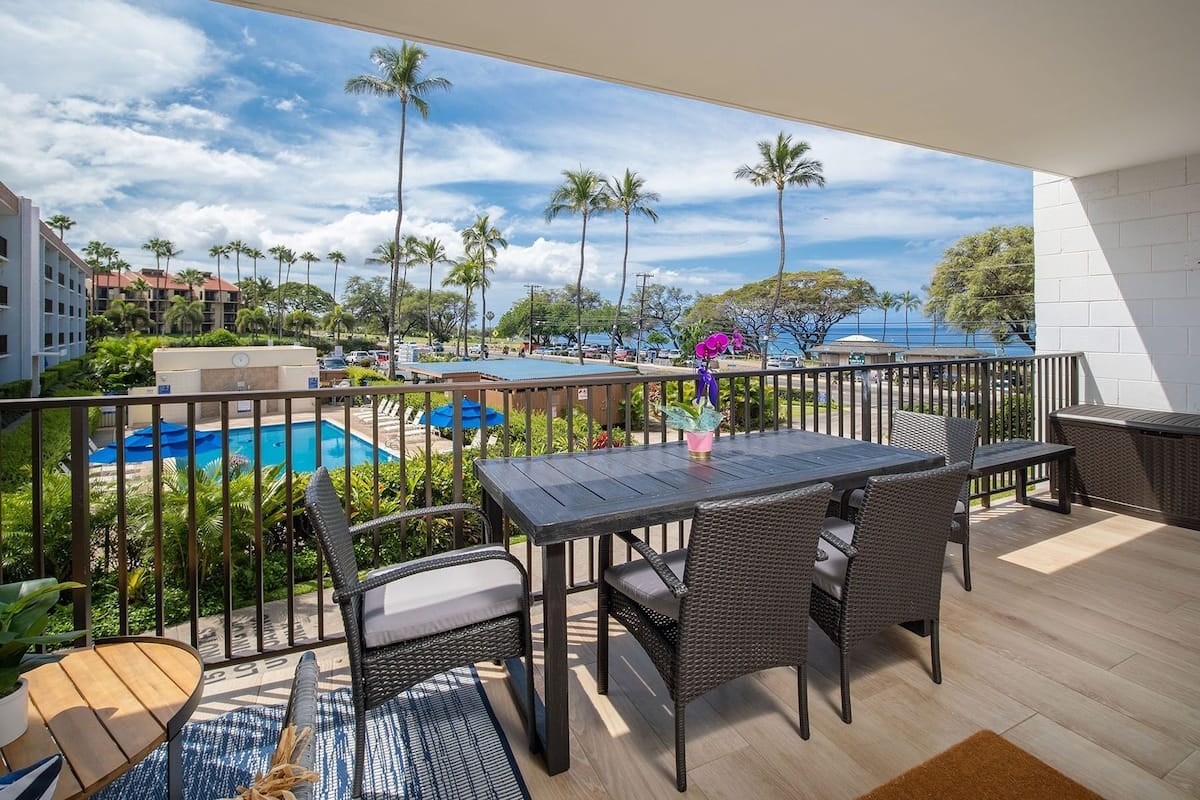A balcony with a pool, wicker furniture, and a view of palm trees and a building in the background.