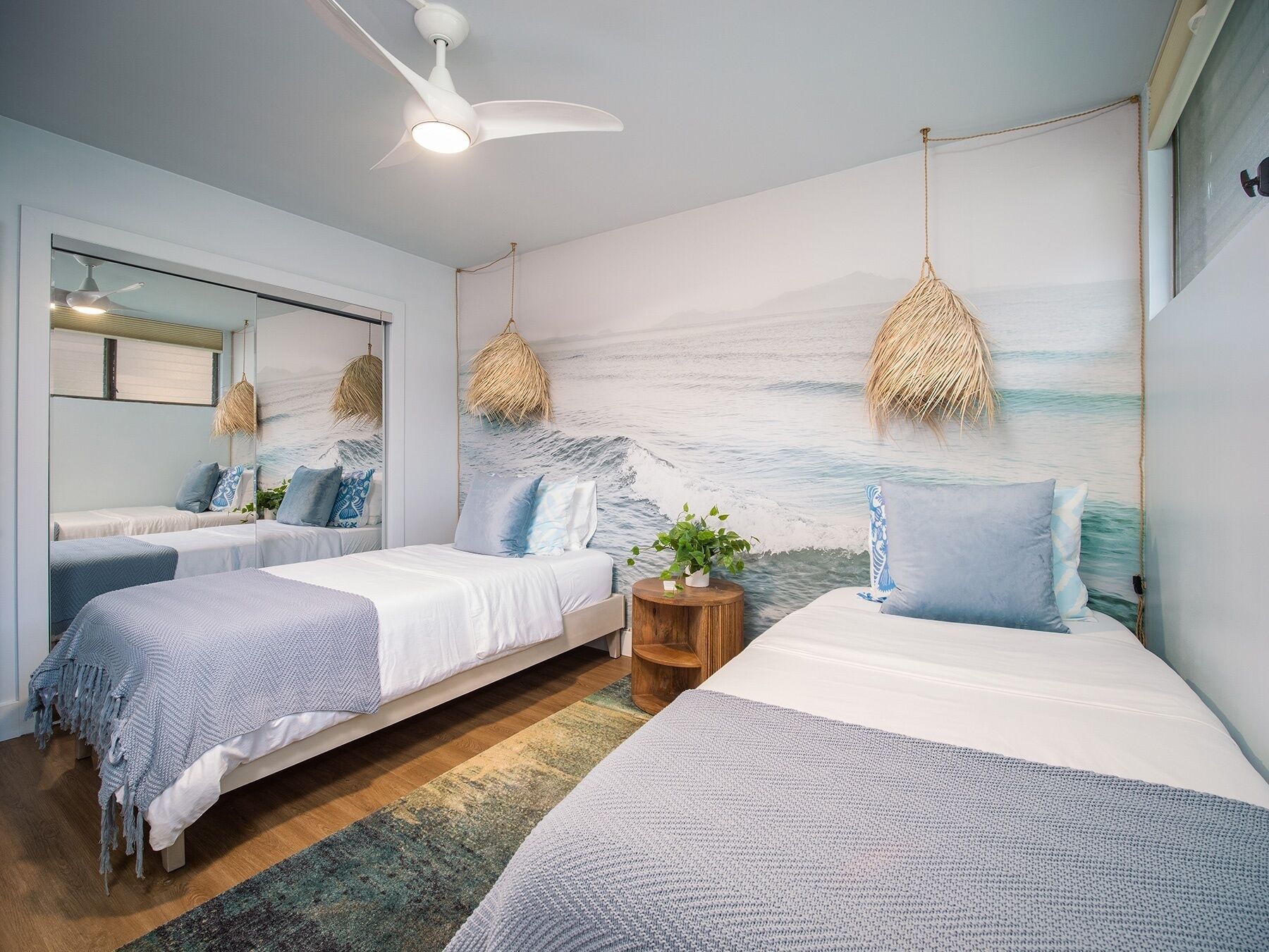Une chambre moderne avec deux lits, un grand miroir, un ventilateur de plafond et une fresque représentant une scène de plage.