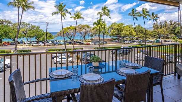 A balcony with a table set for dining, overlooking a parking area and palm trees, with a view of the ocean and a clear sky. - Maui Parkshore by Coldwell Banker Island Vacations (Kihei)