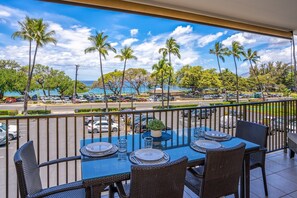 A balcony with a table set for dining, overlooking a parking area and palm trees, with a view of the ocean and a clear sky.