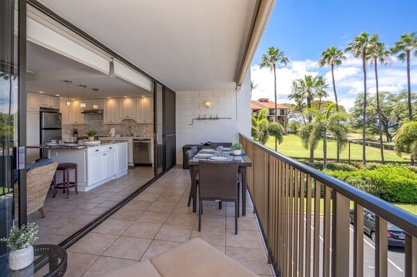 A balcony with a dining table, chairs, and a view of a lush garden with palm trees.