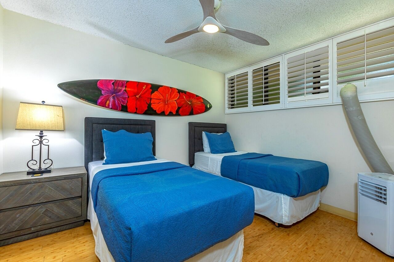 A room with two beds, a ceiling fan, a wall-mounted surfboard decoration, and a window with blinds.