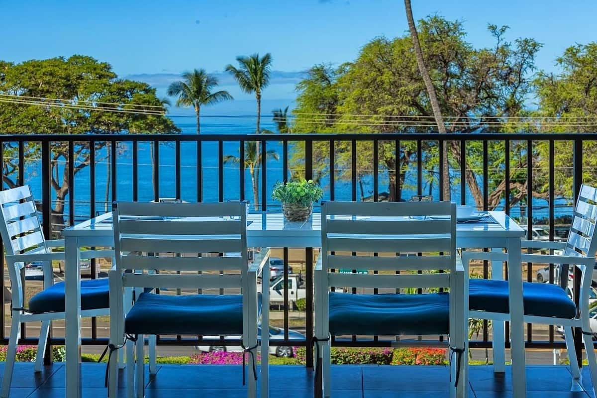 A table with four chairs, a potted plant, and a view of the ocean and palm trees.