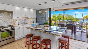 A modern kitchen with white cabinets, a large island with stools, and a view of the ocean. - Maui Parkshore by Coldwell Banker Island Vacations (Kihei)