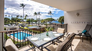 A balcony with a glass table set for dining, overlooking a swimming pool and a parking area with palm trees. - Maui Parkshore by Coldwell Banker Island Vacations (Kihei)