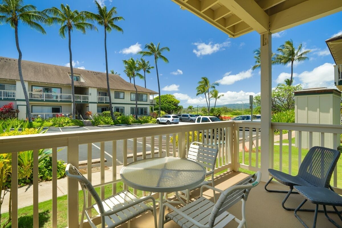 A covered outdoor area with a table and chairs, a view of a building with a balcony, and palm trees.