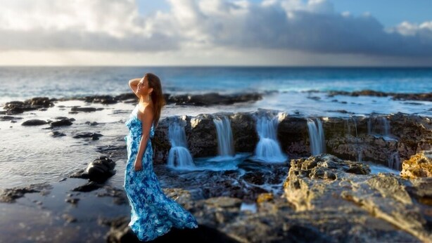 A woman in a blue dress standing by a rocky shoreline with a waterfall. - Kauai Regency at Poipu Kai by Coldwell Banker Island Rentals (Koloa)