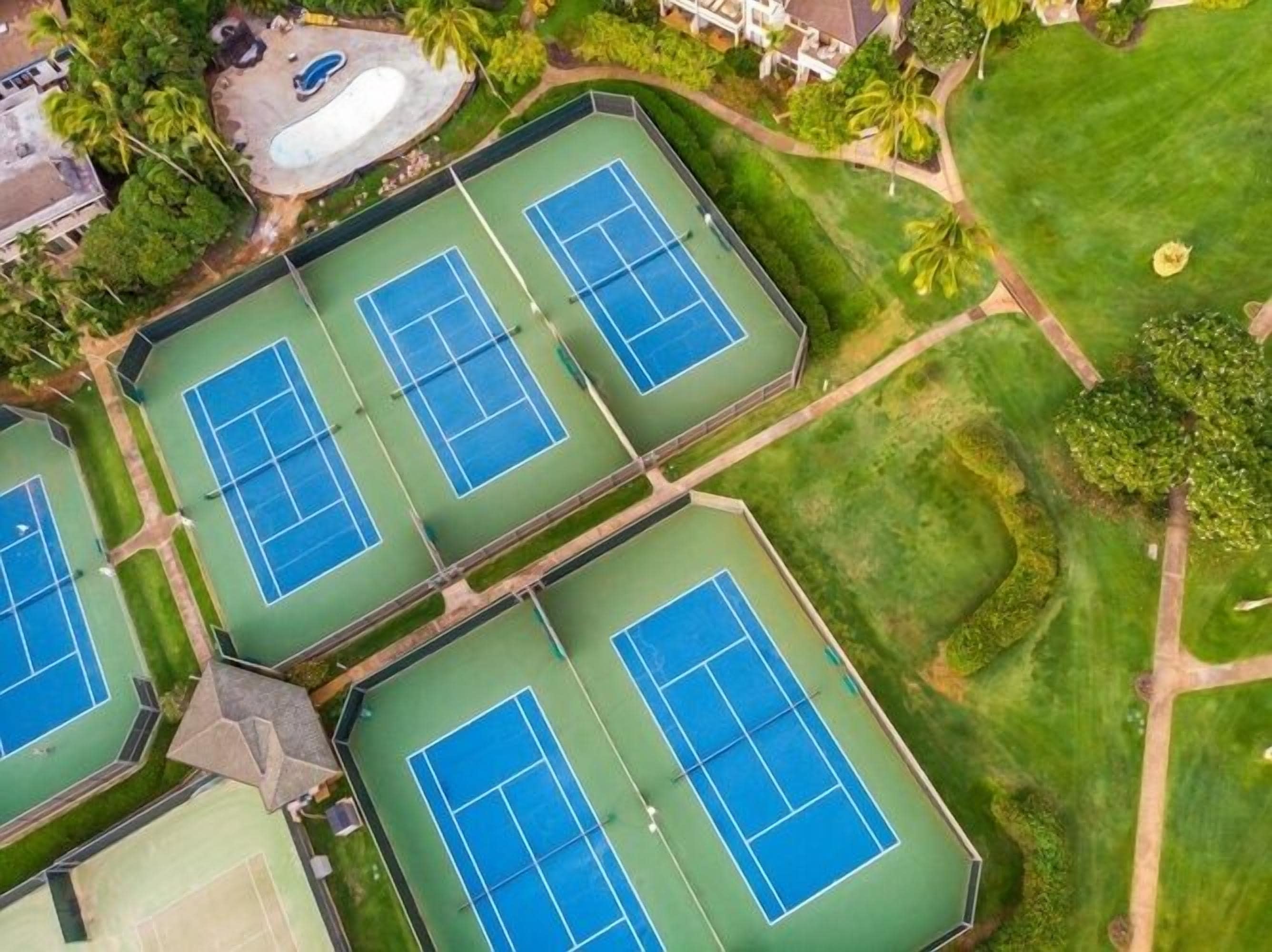 An aerial view of a tennis court complex with multiple courts, surrounded by greenery and a swimming pool.