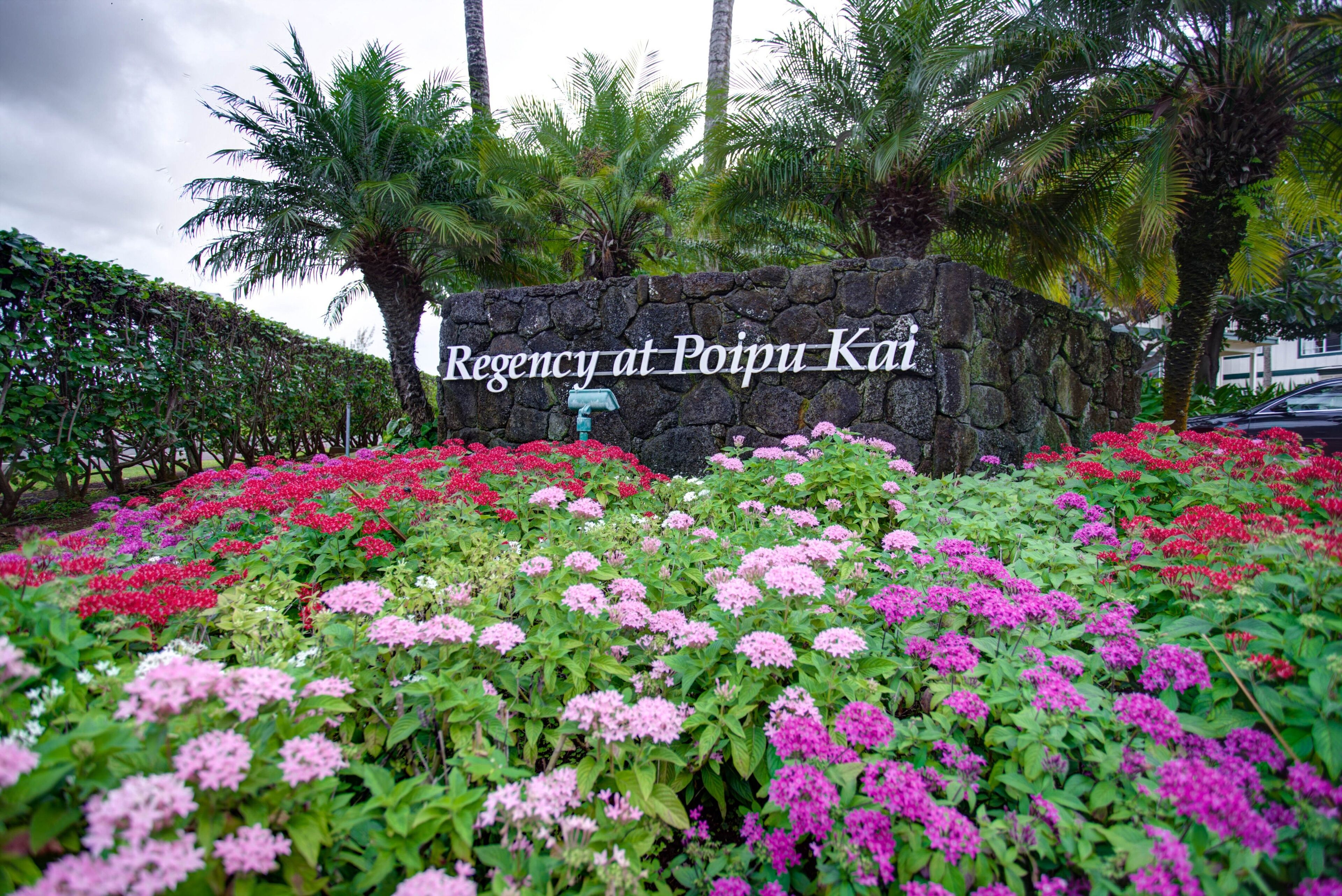 Mural de pedra com uma placa que diz "Regency at Poipu Kai", rodeada por canteiros de flores vibrantes.