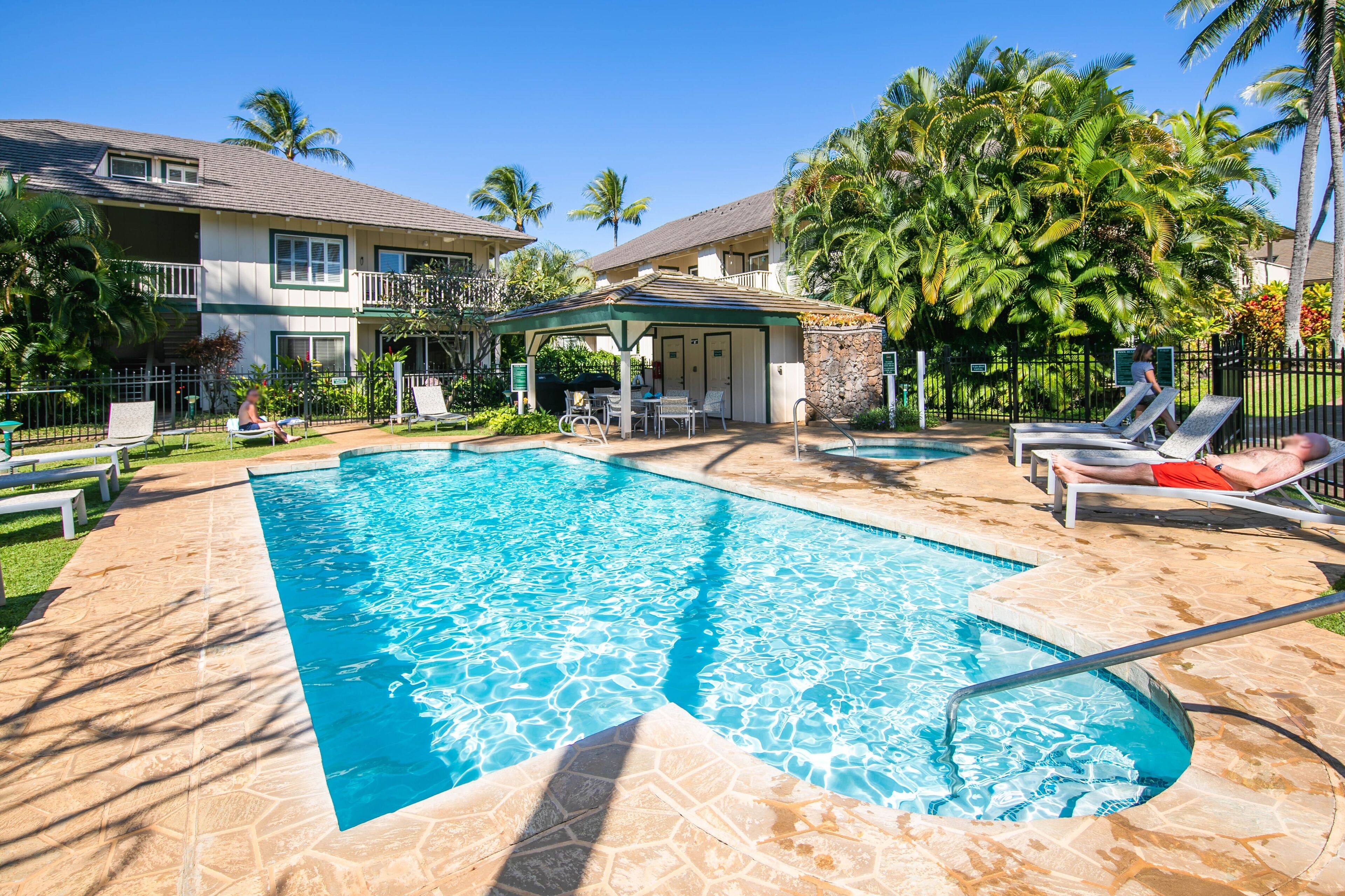 A swimming pool with lounge chairs and a covered seating area, surrounded by palm trees and a two-story building.