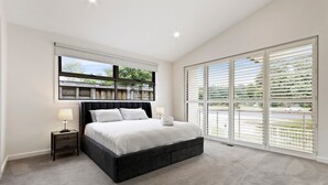 A modern bedroom with a large bed, a nightstand, a wall-mounted shelf, and a large window with blinds.