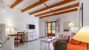 Ein helles Wohnzimmer mit Holzboden, einem an der Wand befestigten Fernseher, einer Essecke mit Tisch und Stühlen und einer gemütlichen Sitzecke mit einer Couch und Kissen.