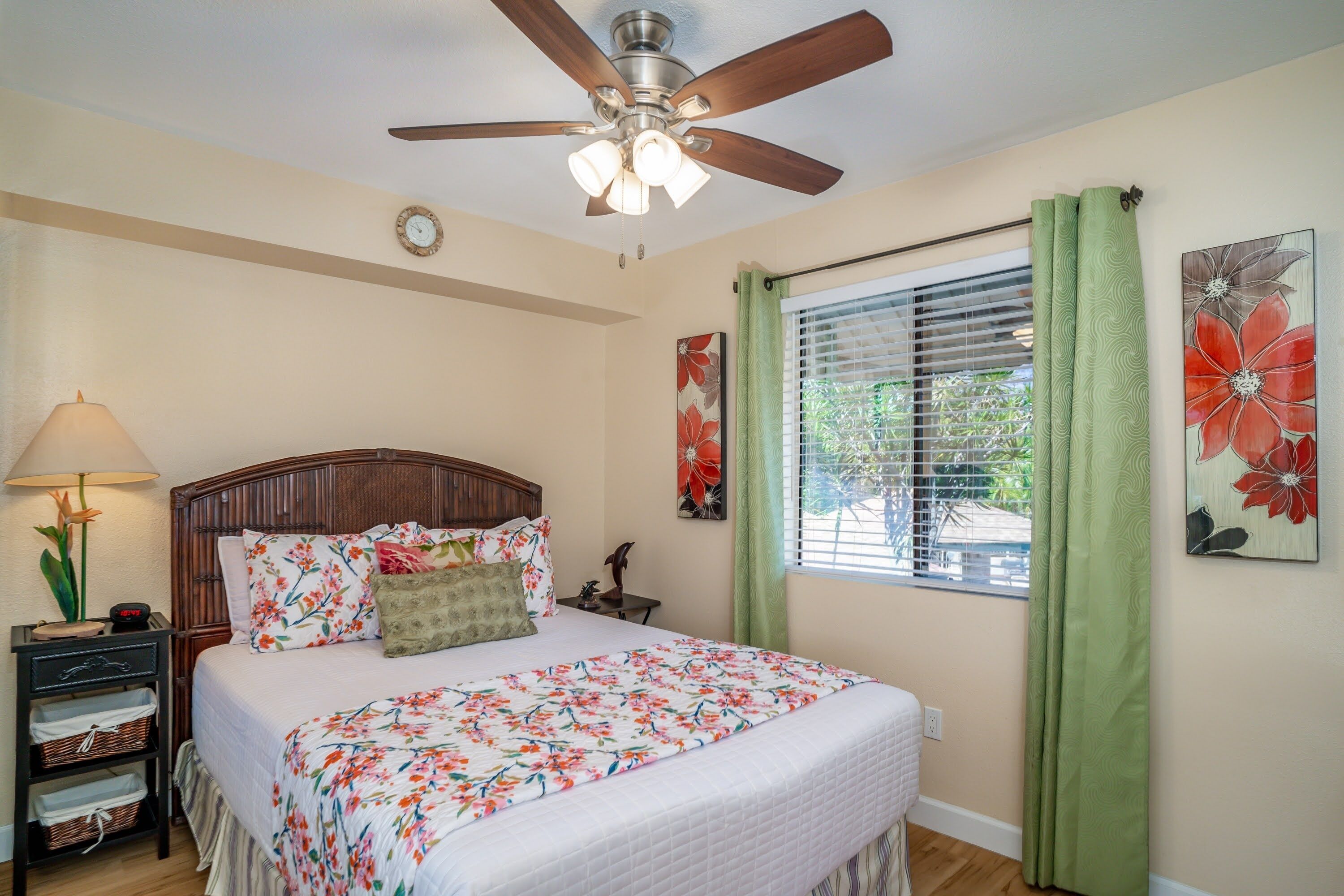 A bedroom with a bed, a nightstand, a lamp, a ceiling fan, a window with curtains, and a wall clock.