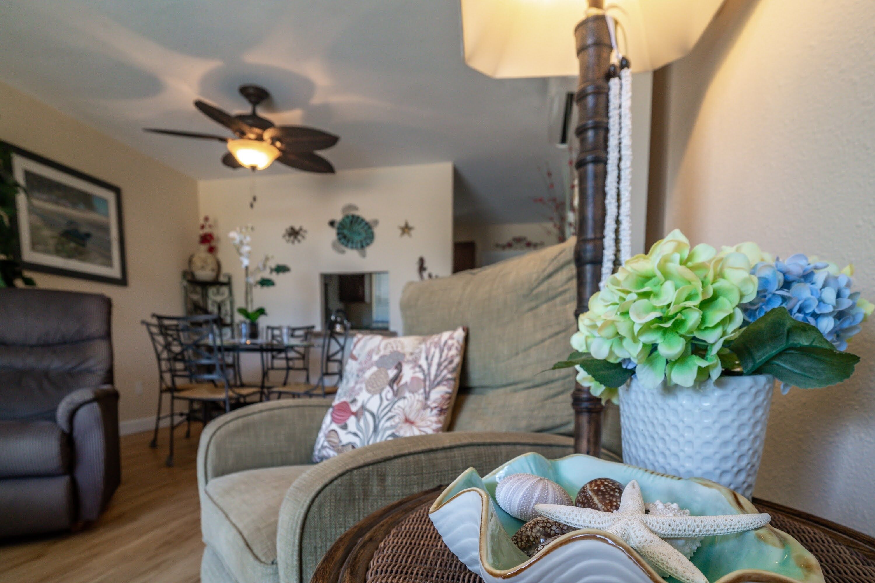 Un salon chaleureux avec un ventilateur de plafond, un coin repas avec des chaises et une table, et une plante décorative posée sur une table d’appoint.