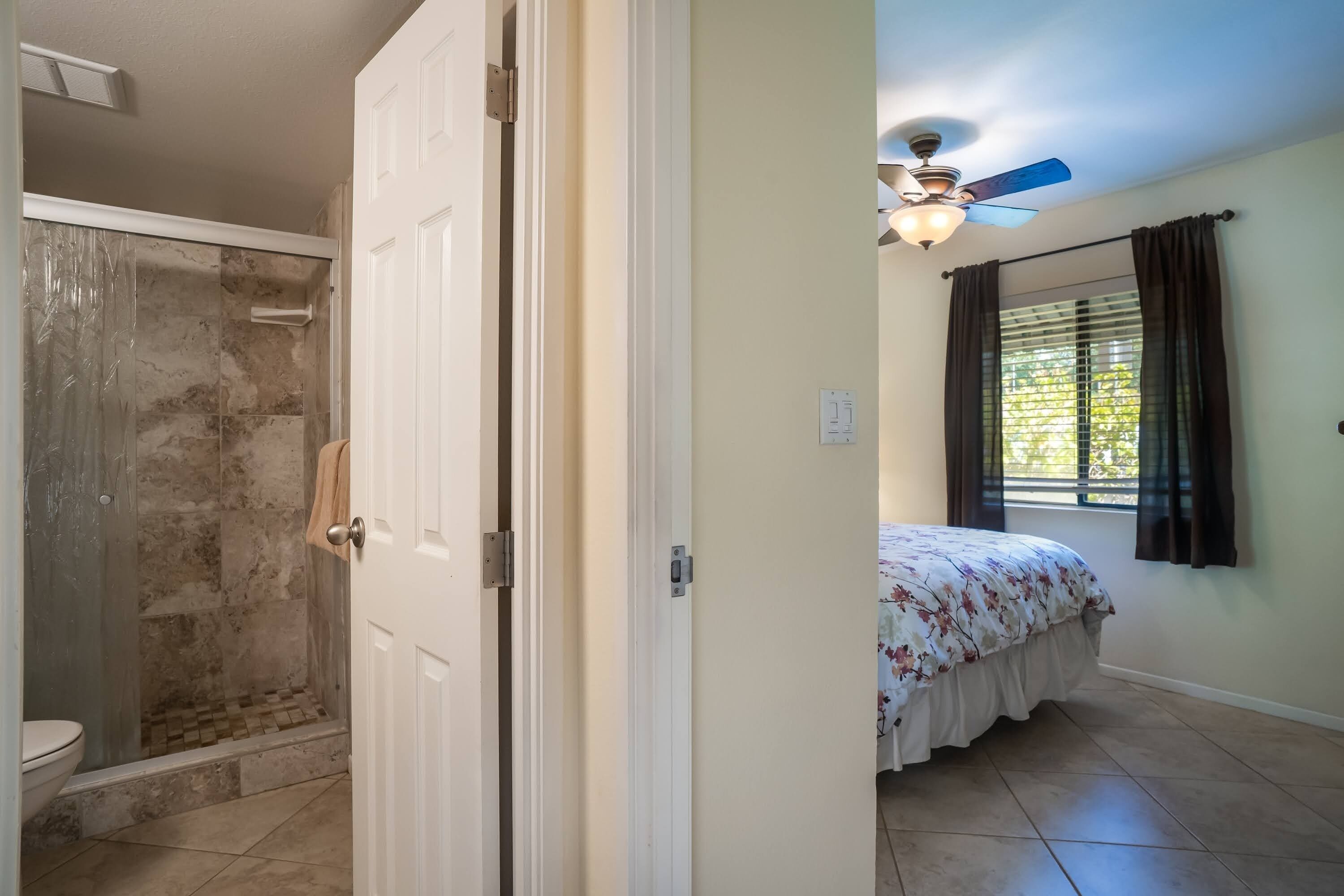 A bathroom with a shower, toilet, and a door leading to a bedroom with a bed and a ceiling fan.